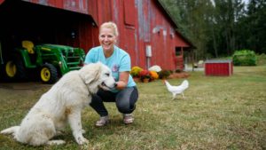 Woman smiling with her hand out to dog. In the background, a big, red barn, tractor and chicken.